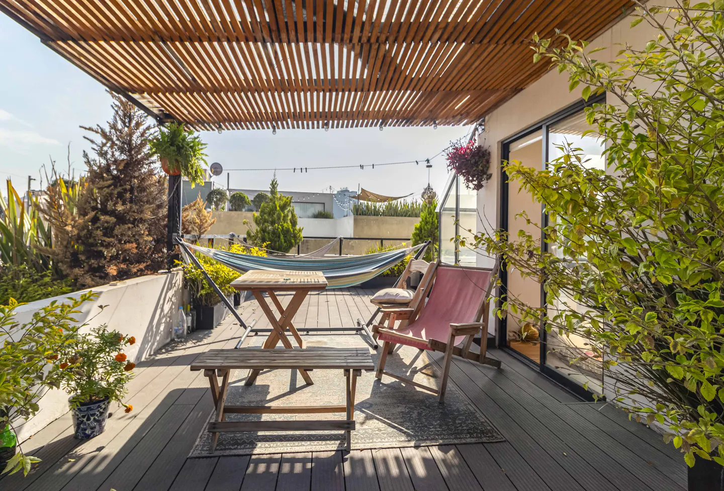 Outdoor patio with wooden pergola, hammock, table, bench, and pink chairs. Green plants and trees surround the space.