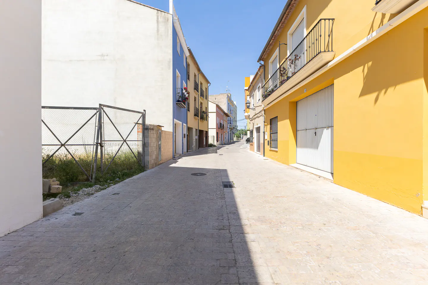 A sunny street view with colorful buildings. Yellow and blue buildings line the street, with a metal fence on the left.