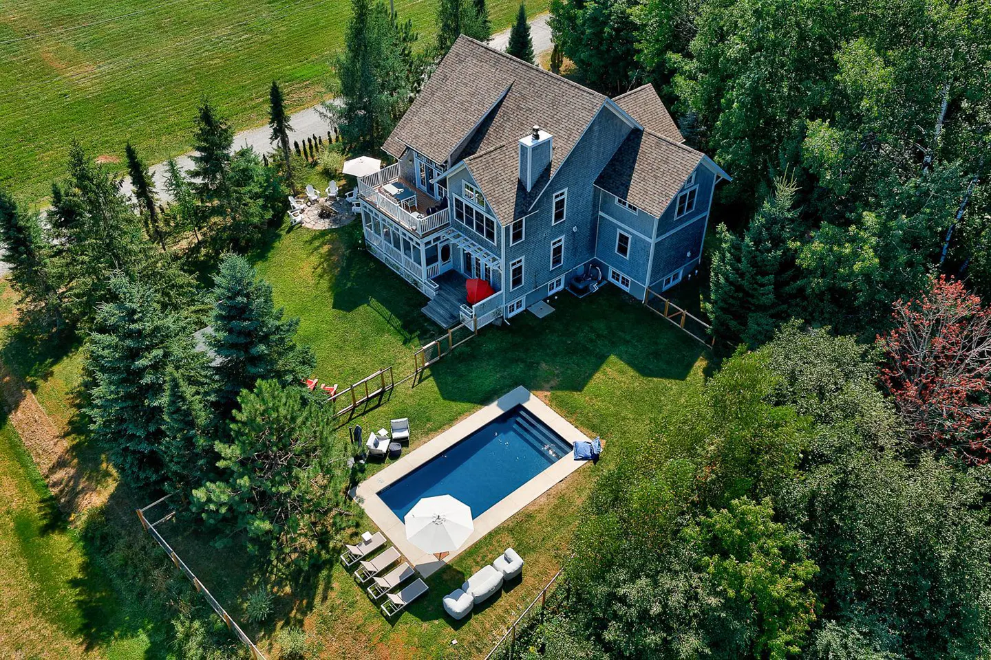 Aerial view of a large, gray house with a pool, surrounded by green grass and trees. Lounge chairs and an umbrella are near the pool.