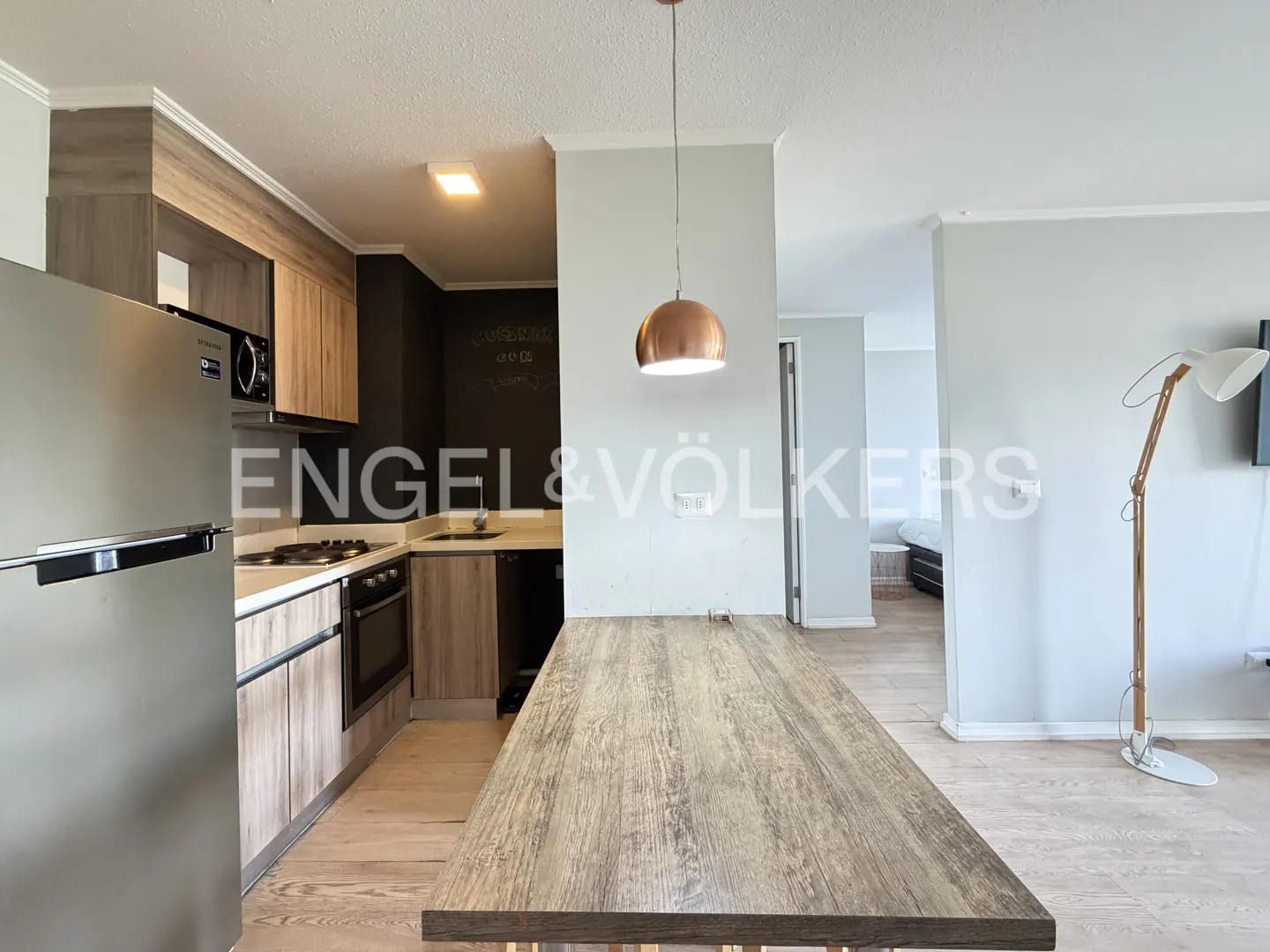 A modern kitchen with stainless steel appliances, wood cabinets, and a long wooden table under a copper pendant light.