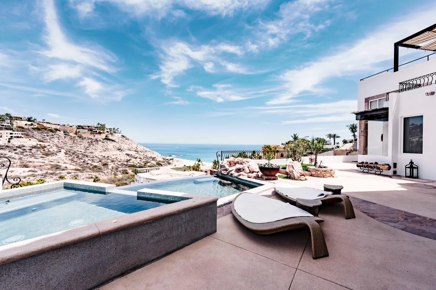 Luxury villa patio with pool, lounge chairs, and ocean view. White building, blue sky, and hillside homes in the background.