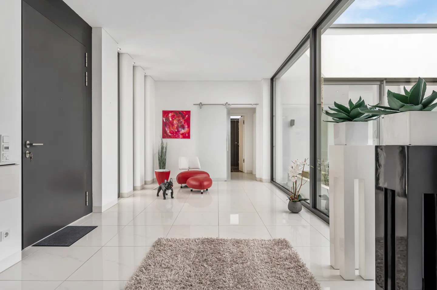 Bright, modern entryway with white tile floors, a gray door, and a large window. A small dog stands near a red chair and ottoman. A beige rug is in the foreground.