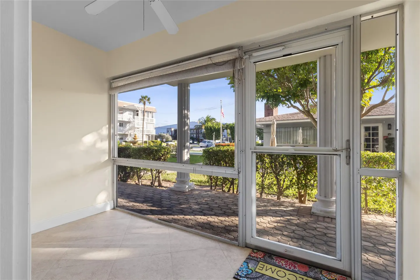 Sunroom view with white framed windows and door, showing a brick patio, manicured hedges, and a building across the street. A welcome mat is on the floor.