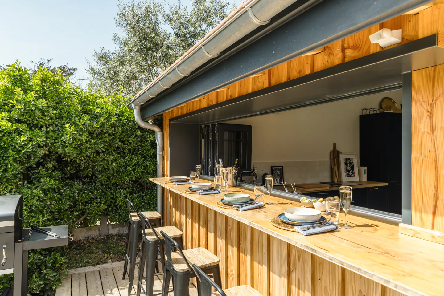 Outdoor kitchen with a wooden bar, stools, and table settings. A grill sits on a wooden deck next to a green hedge.