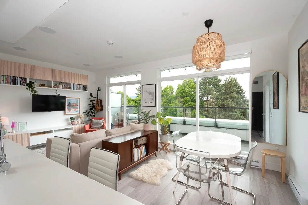 Modern open-plan living room with a beige sofa, wooden sideboard, white dining table, and large windows overlooking trees.