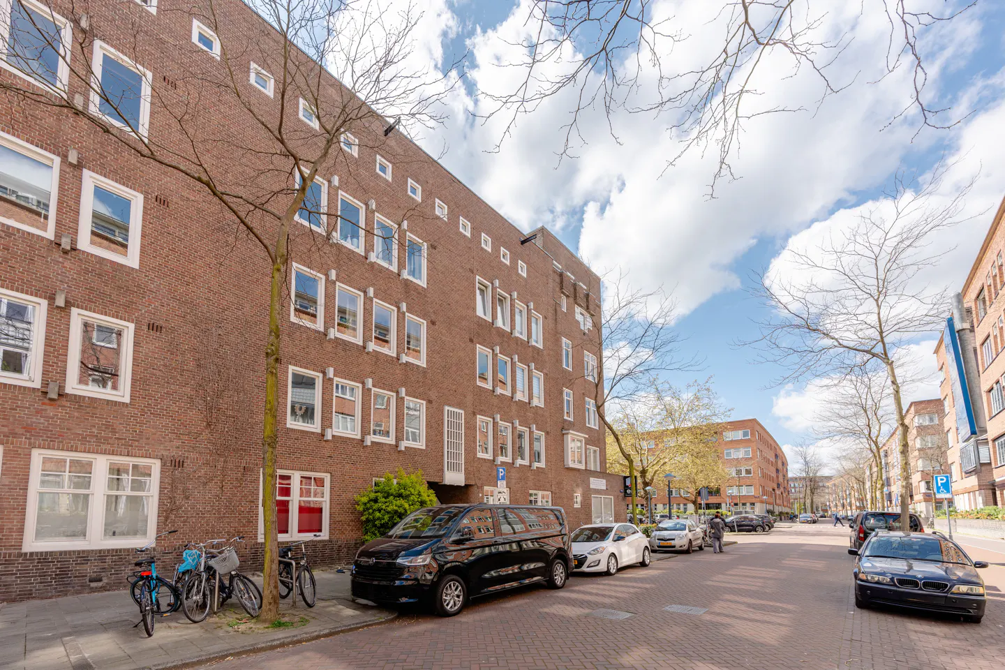 Brick apartment building with white-framed windows on a street with parked cars and bicycles under a partly cloudy sky.