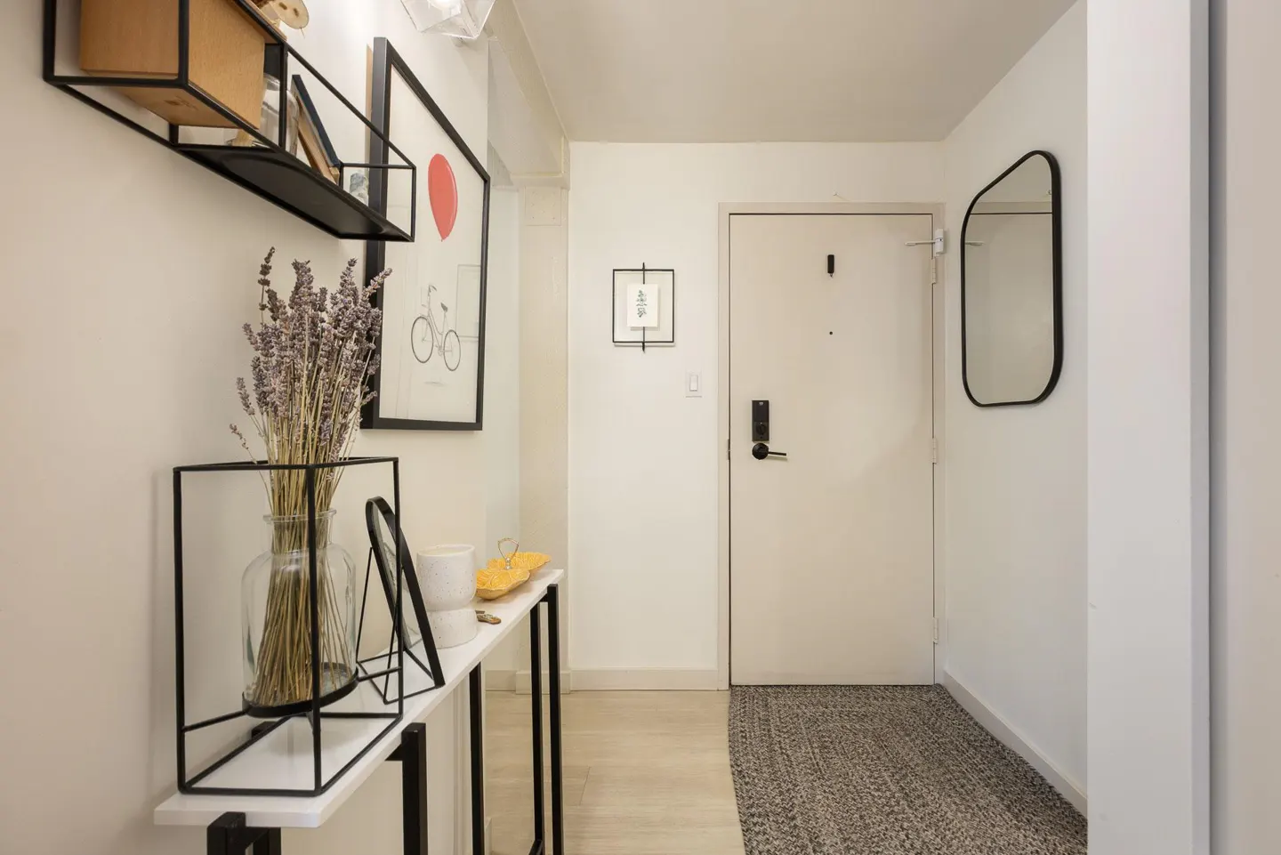 Entryway with white walls, a white door, and a gray rug. A console table with lavender in a vase and framed art adds decor.