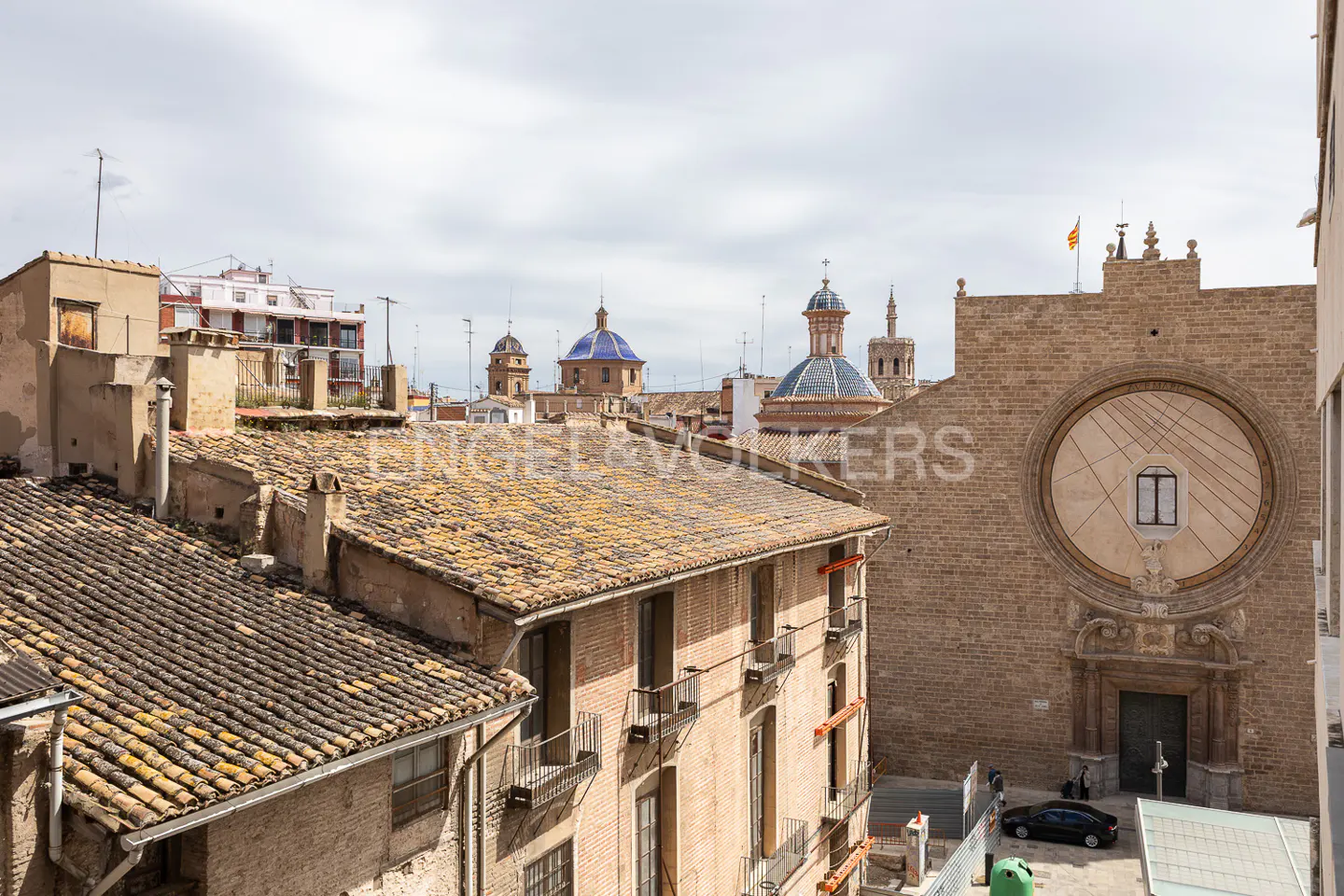 Cityscape view of Valencia, Spain, featuring tiled rooftops, church domes, and a sundial facade on a brick building.