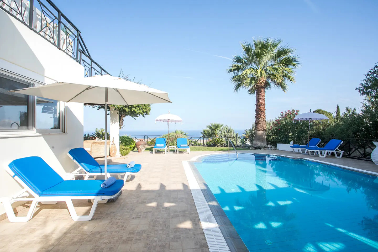 Outdoor pool with blue lounge chairs, white umbrellas, and a palm tree against a clear blue sky.