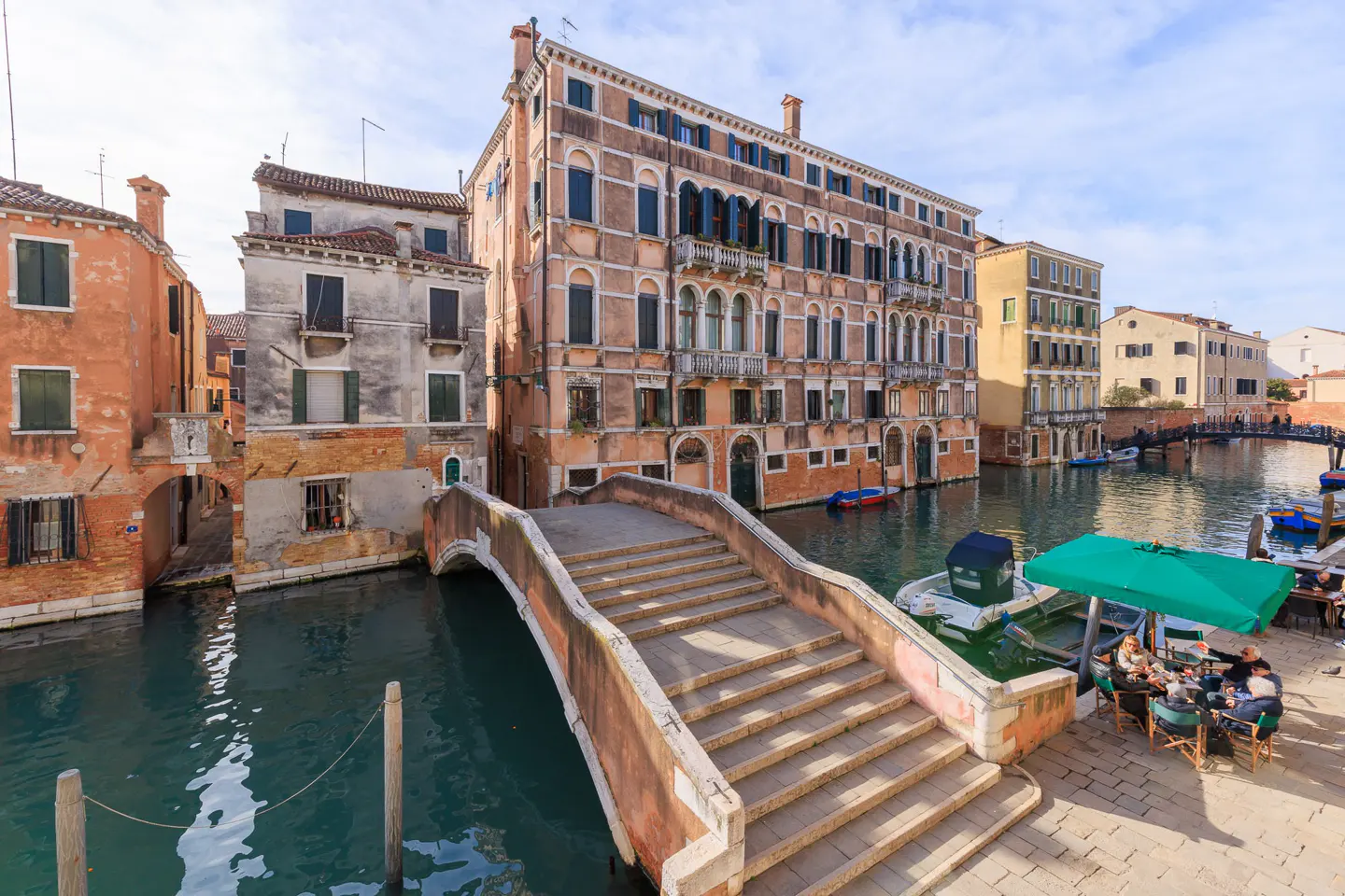 Venice canal view with a stone bridge, buildings, and people dining under a green umbrella.