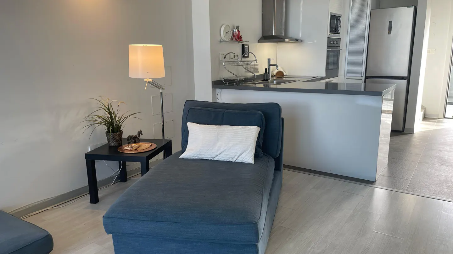 Living room with a blue chaise lounge, white pillow, black side table, lamp, and open kitchen in the background.