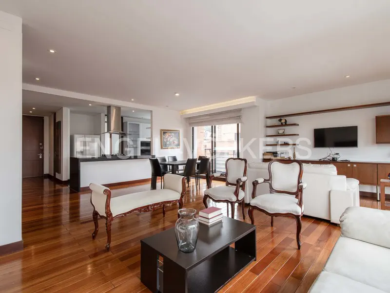 Open-concept living space with hardwood floors, white sofas, chairs, and a dark wood coffee table. Kitchen and dining area in the background.
