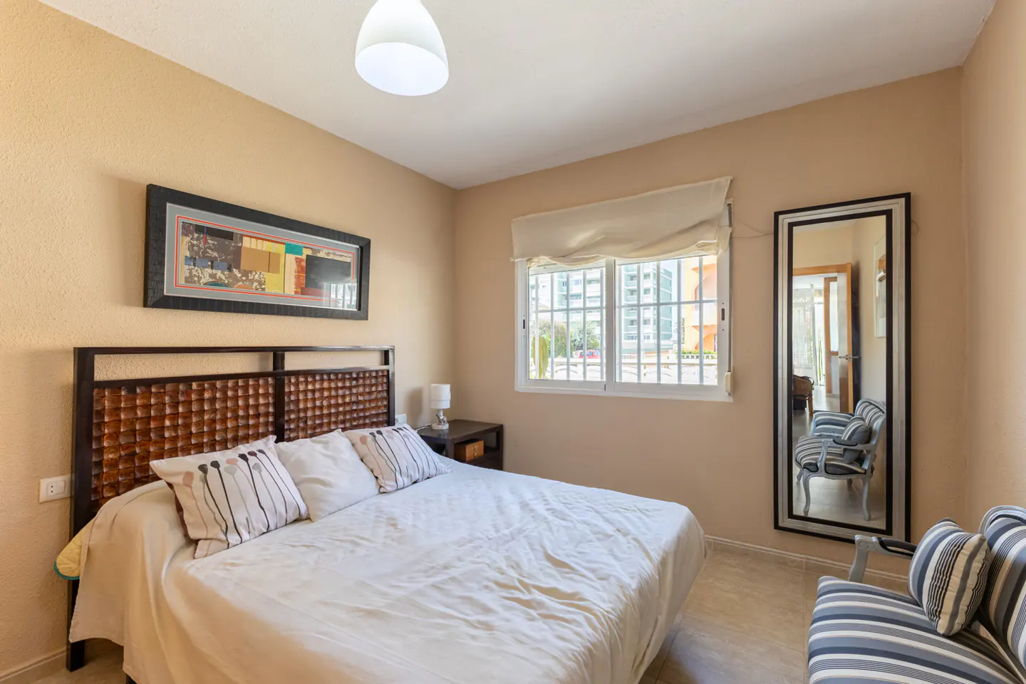 Bedroom with tan walls, a bed with white linens, a woven headboard, and a window with white blinds. A mirror reflects a striped chair.