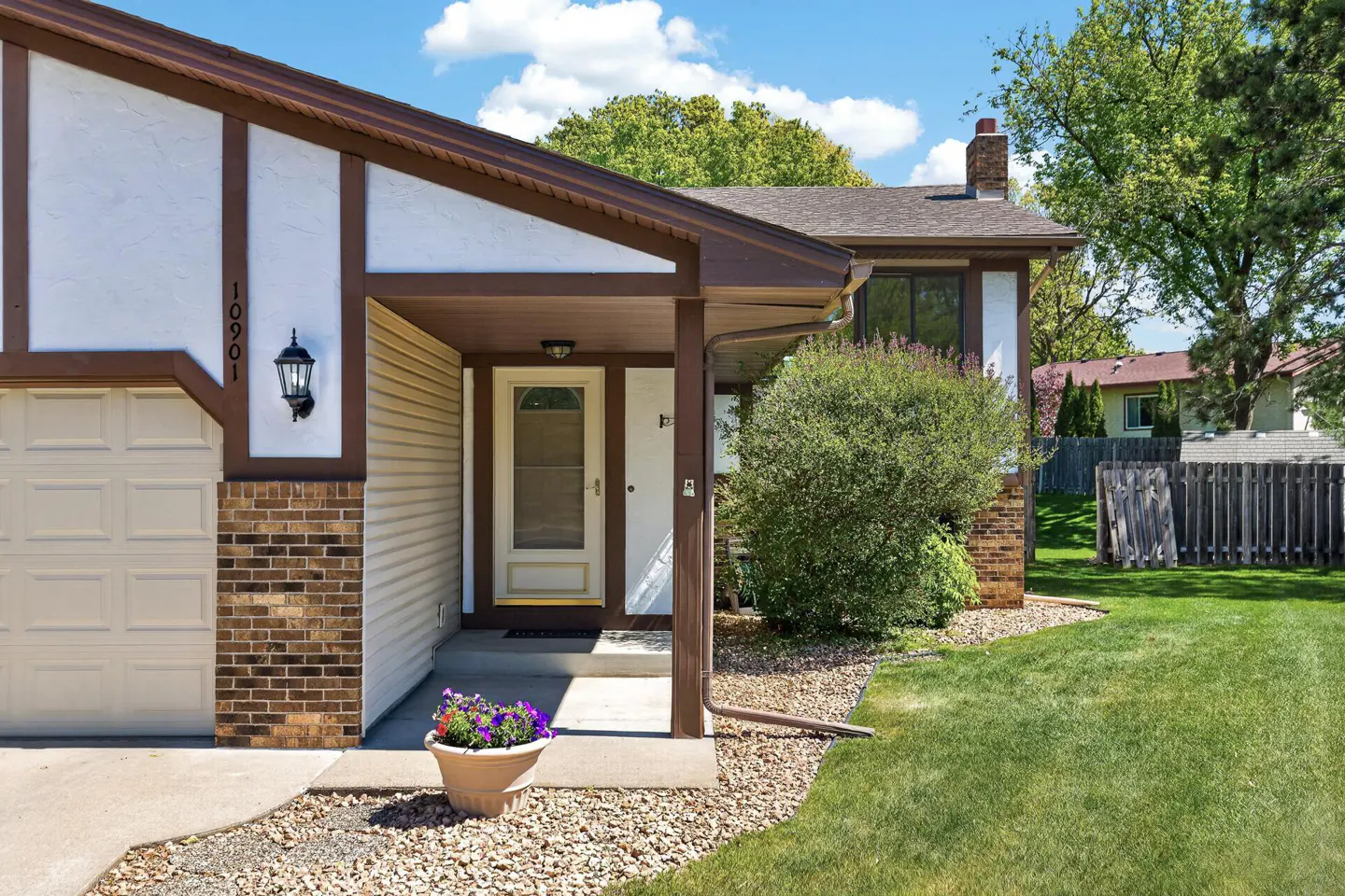 Exterior of a two-story house with brown trim, a white door, and a green lawn. A flower pot sits near the walkway.