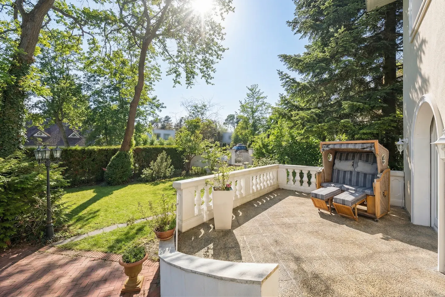Sunny patio with a wicker beach chair, footrests, and a white balustrade overlooking a green lawn and trees.