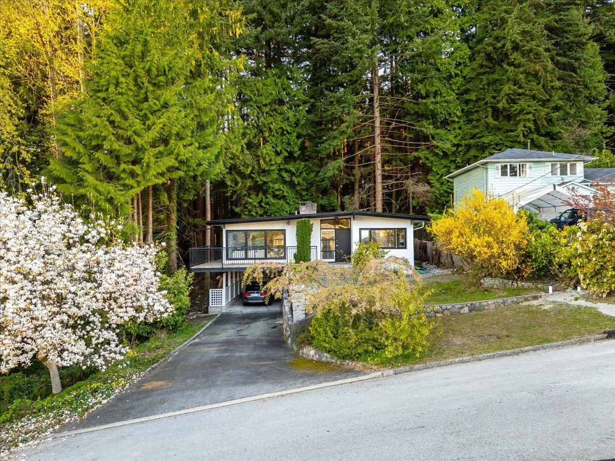 Modern white house with black trim, surrounded by lush green trees and flowering bushes. A car is parked under the elevated structure.