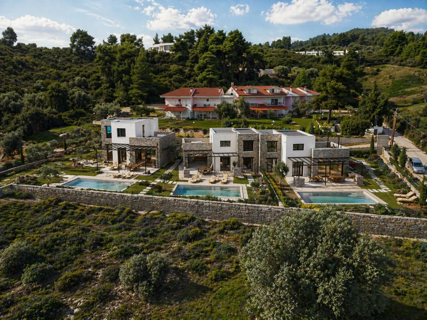Modern white villas with stone accents and private pools on a green hillside under a blue sky with scattered clouds.