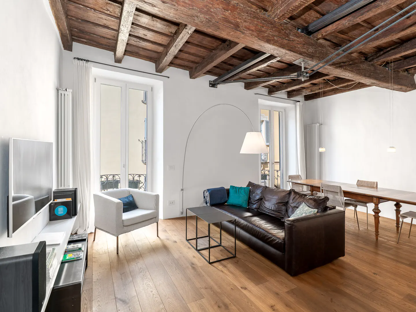 Living room with wood floors, white walls, and exposed wood beam ceiling. A brown leather sofa and white chair sit near windows.