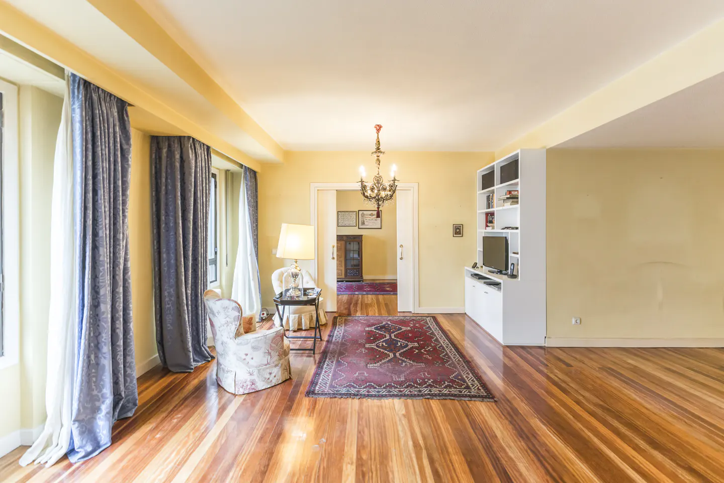 Living room with hardwood floors, yellow walls, and a red patterned rug. A white bookcase and TV stand are on the right. Curtains frame the windows.