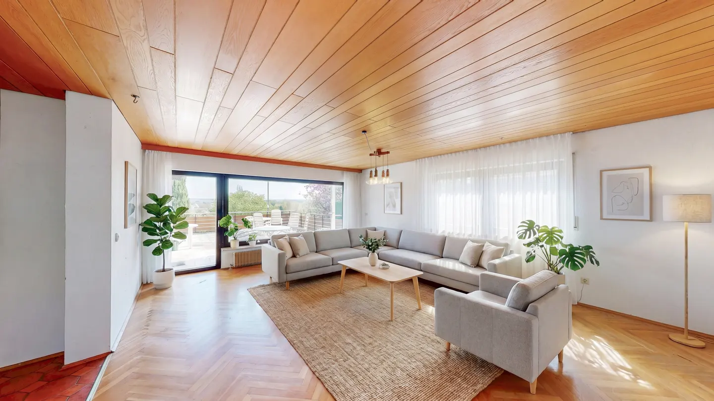 Bright living room with wood ceiling, herringbone floors, and a large gray sectional sofa. A jute rug sits under a light wood coffee table. Large windows offer a view of the outdoors.