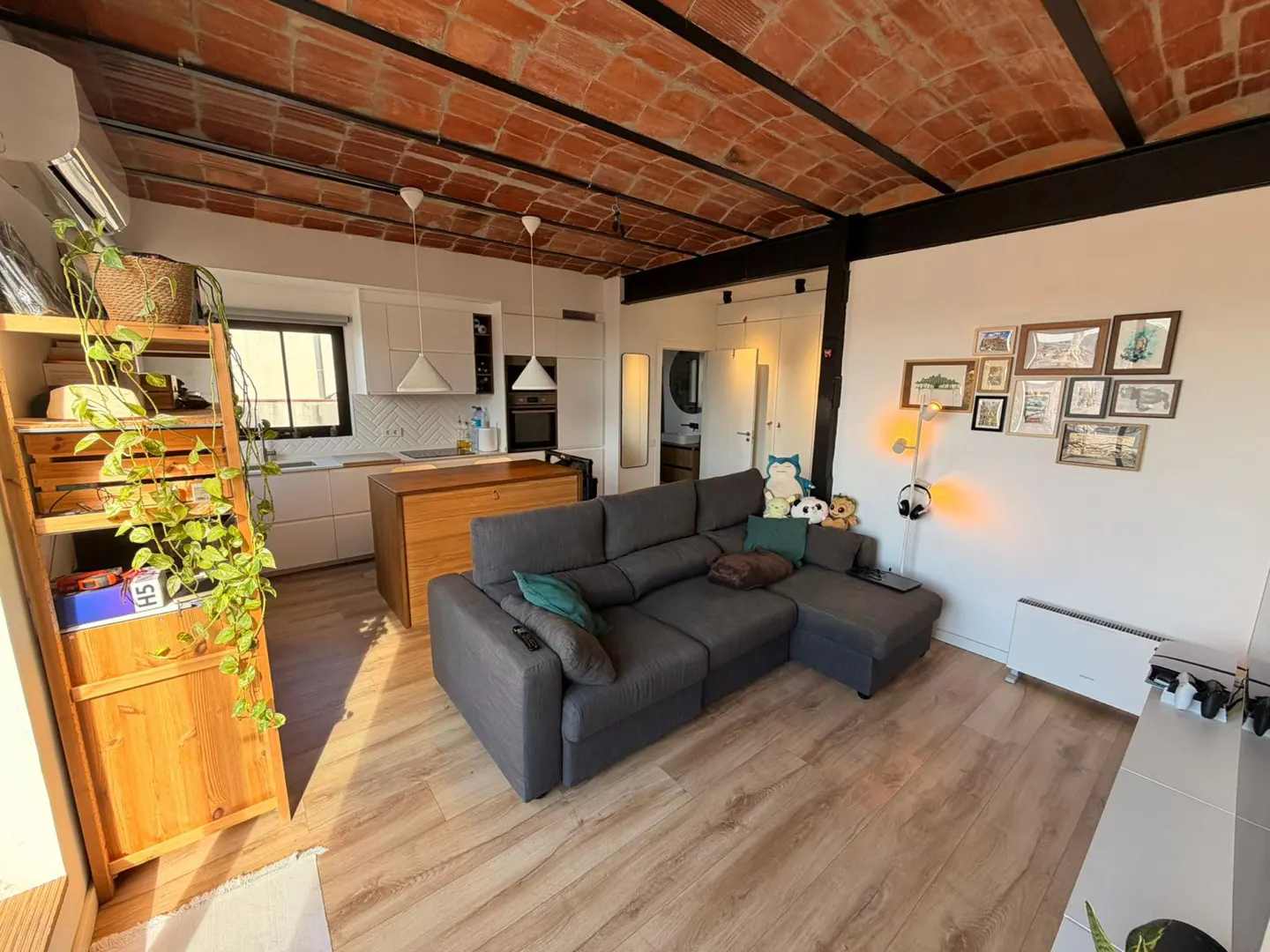 Open-concept apartment with a gray sectional sofa, wood floors, and a brick ceiling. A kitchen island and white cabinets are visible.