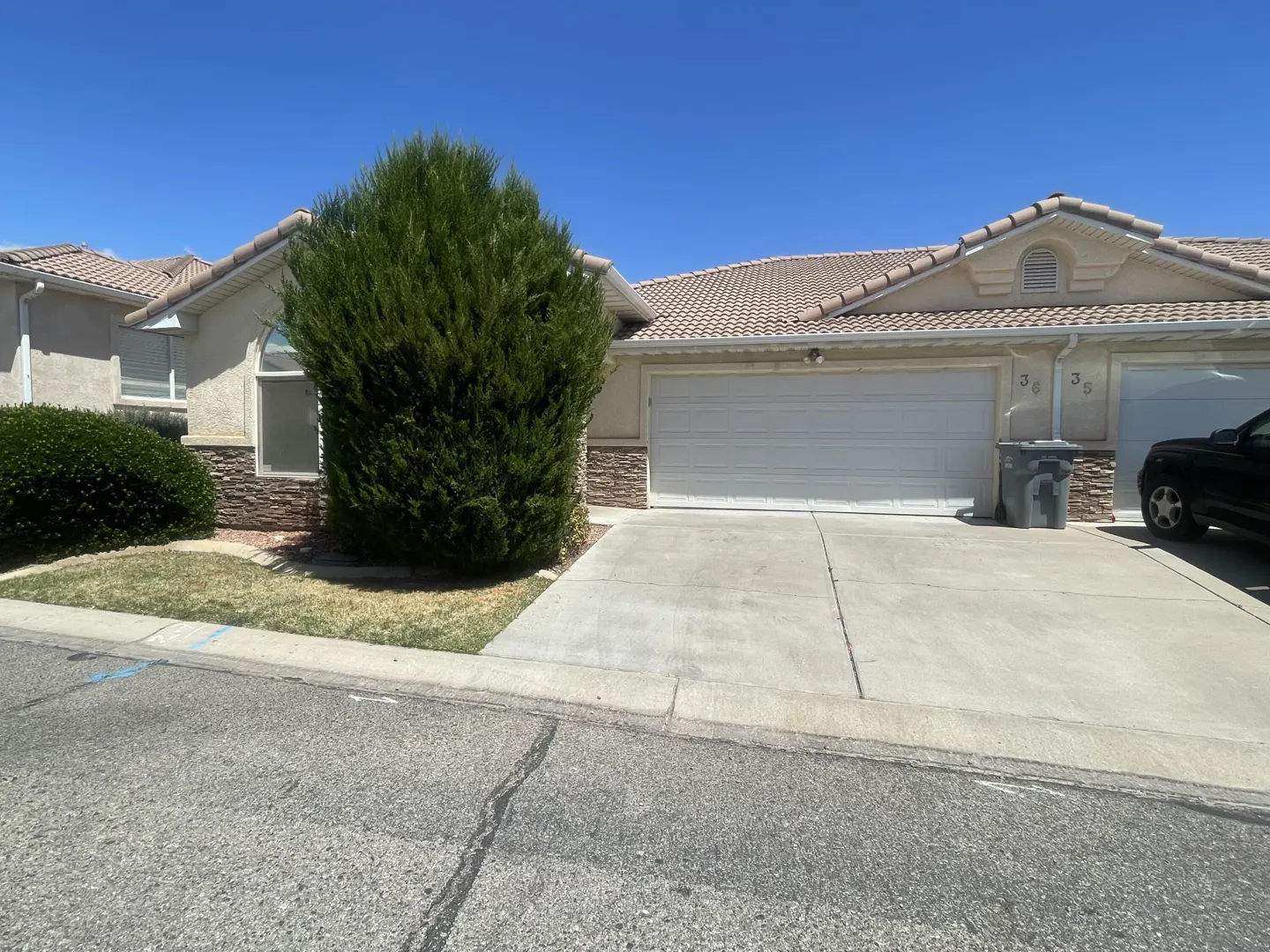 Beige townhome with a white garage door, stone accents, and a green bush. A black truck is parked in the driveway. Blue sky.