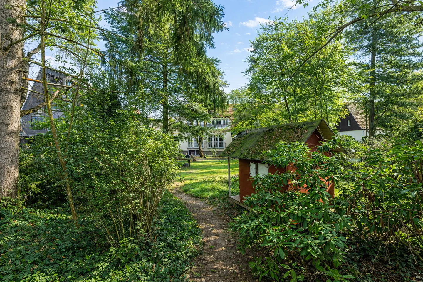 View of a white house through trees, with a red shed on the right and a path leading to the house.