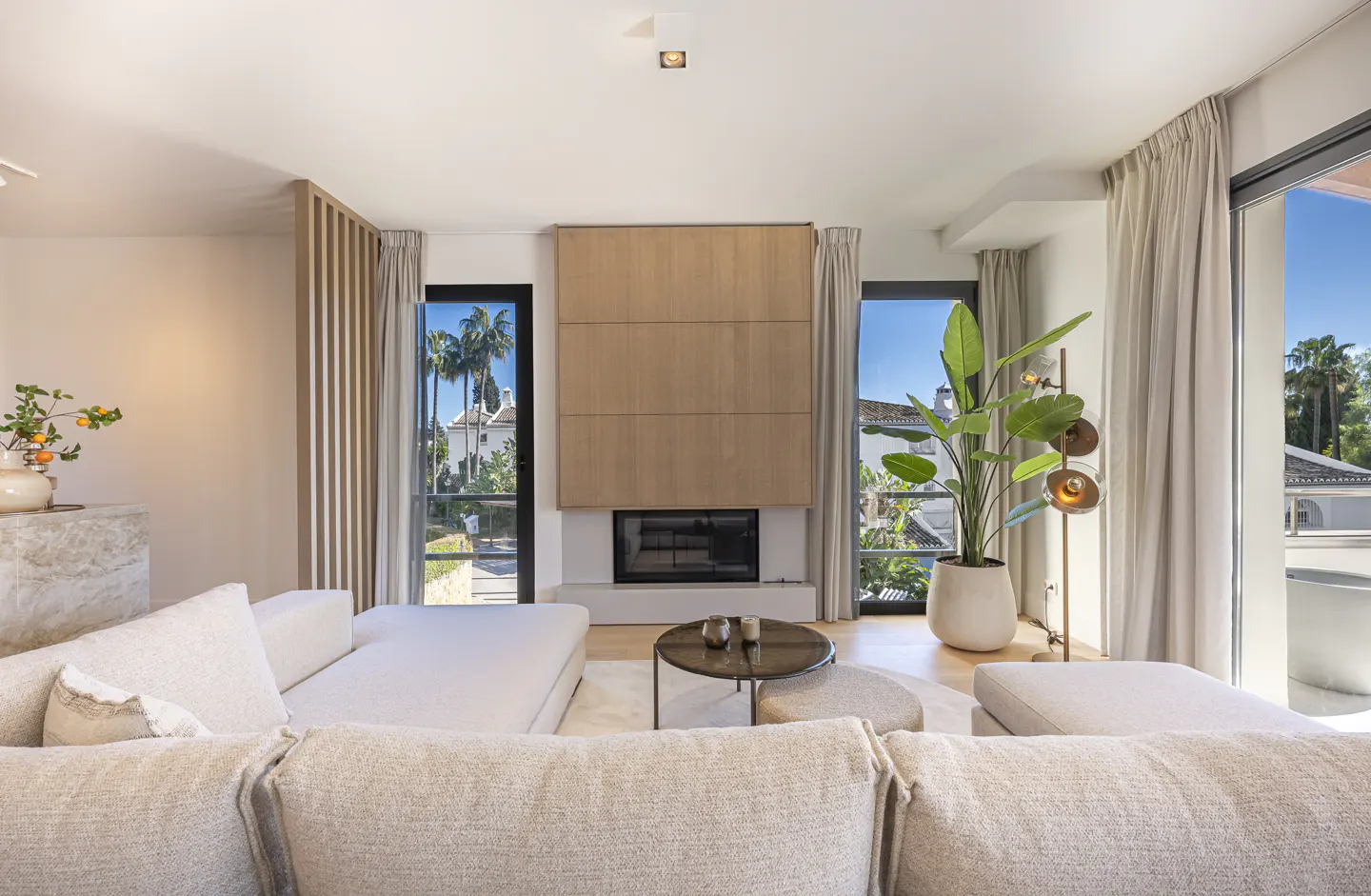 Modern living room with a beige sectional sofa, central fireplace, large potted plant, and floor-to-ceiling windows.
