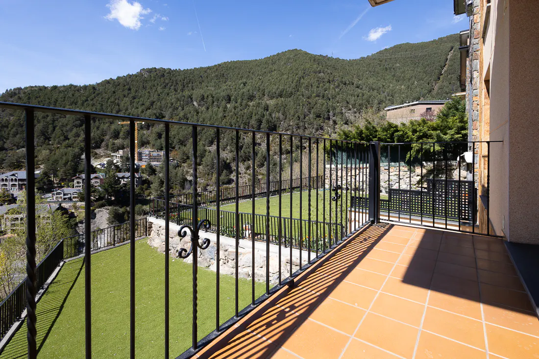 Balcony view with black iron railing, terracotta tiles, and green lawn. Mountain and blue sky background.