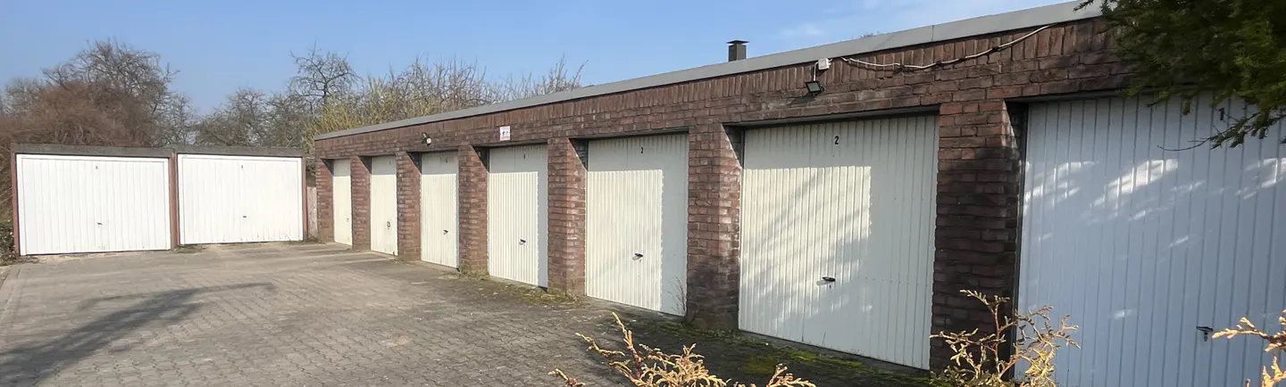 Row of garages with white doors and brick walls, numbered, on a paved driveway under a blue sky.