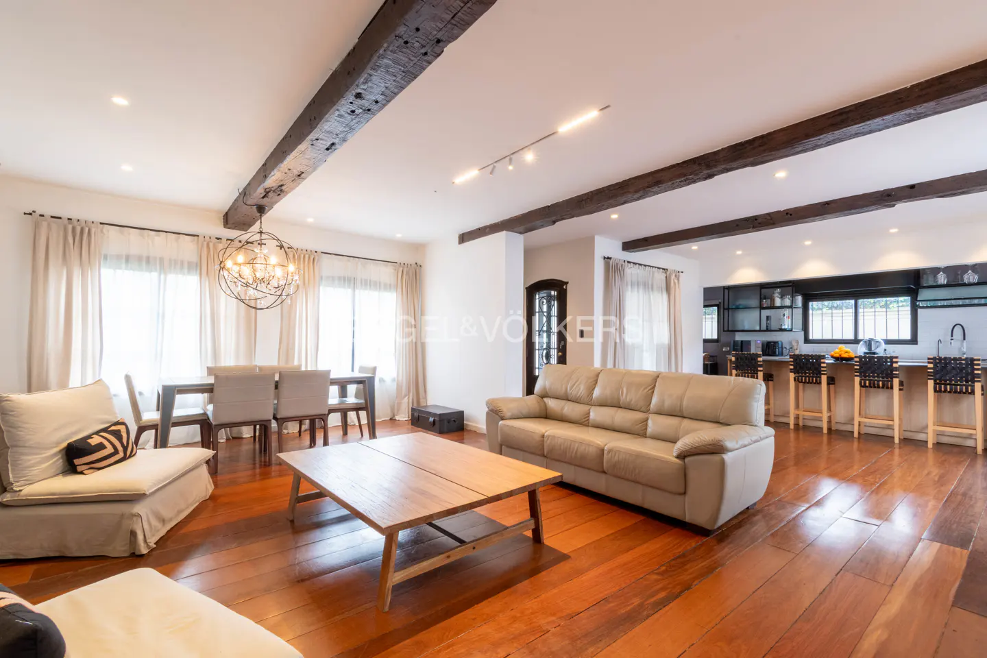 Open-concept living area with hardwood floors, exposed wood beams, beige sofa, wooden coffee table, and dining space.