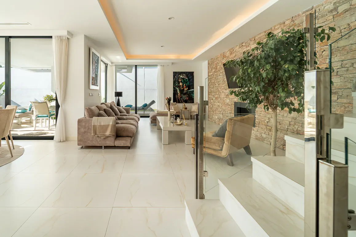 Bright, modern living room with a brown sofa, white tile floors, and a stone accent wall with a tree. Glass staircase in the foreground.