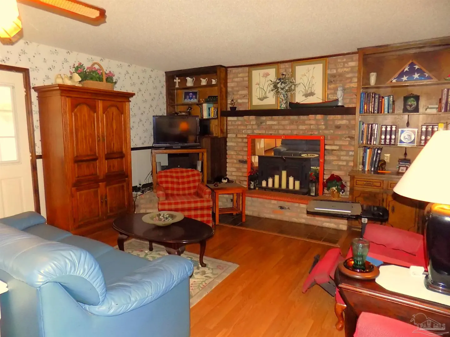 A living room with a blue sofa, wooden floors, and a brick fireplace with candles. There is a TV, bookshelves, and floral wallpaper.