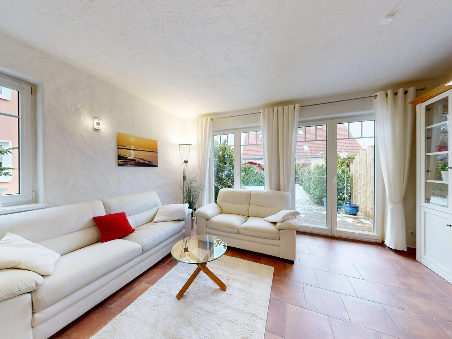 Living room with white sofas, glass table, and white rug on a terracotta tile floor. Sunlight streams through the patio doors.