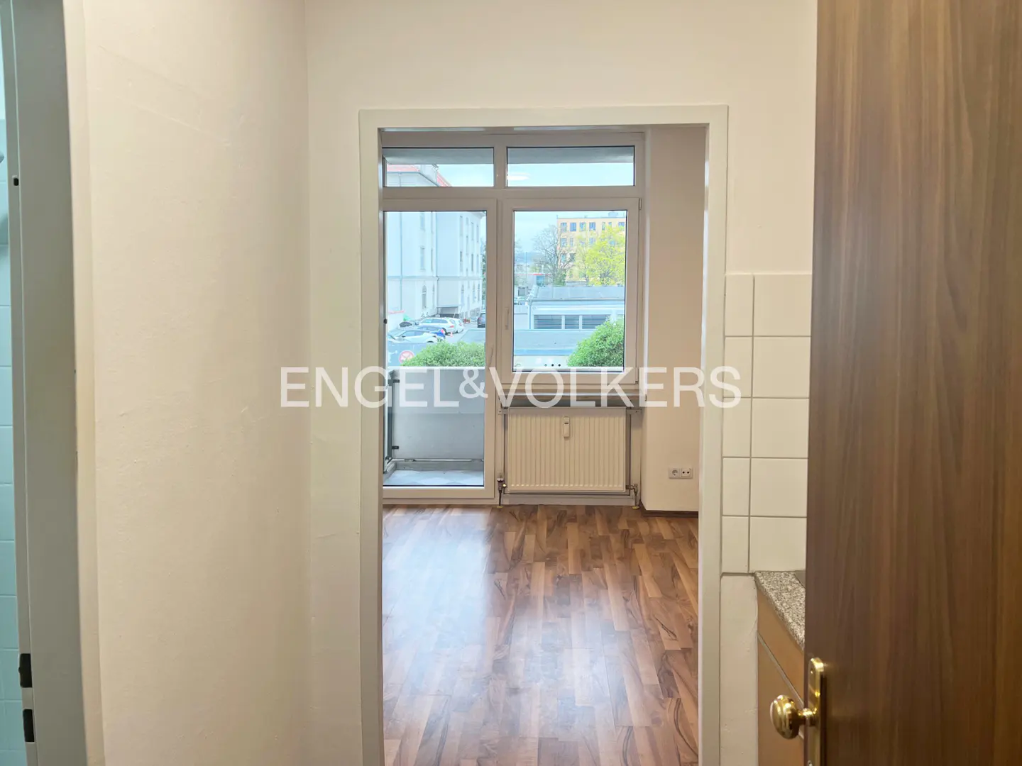 View from a doorway into a room with wood floors and a balcony. Outside, buildings and cars are visible.