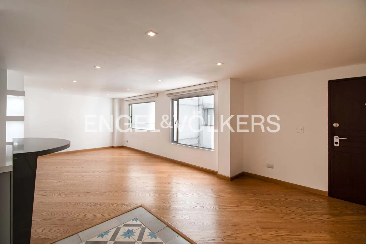 Bright, empty room with hardwood floors, white walls, and two windows. A dark brown door is on the right. Engel & Volkers logo is visible.