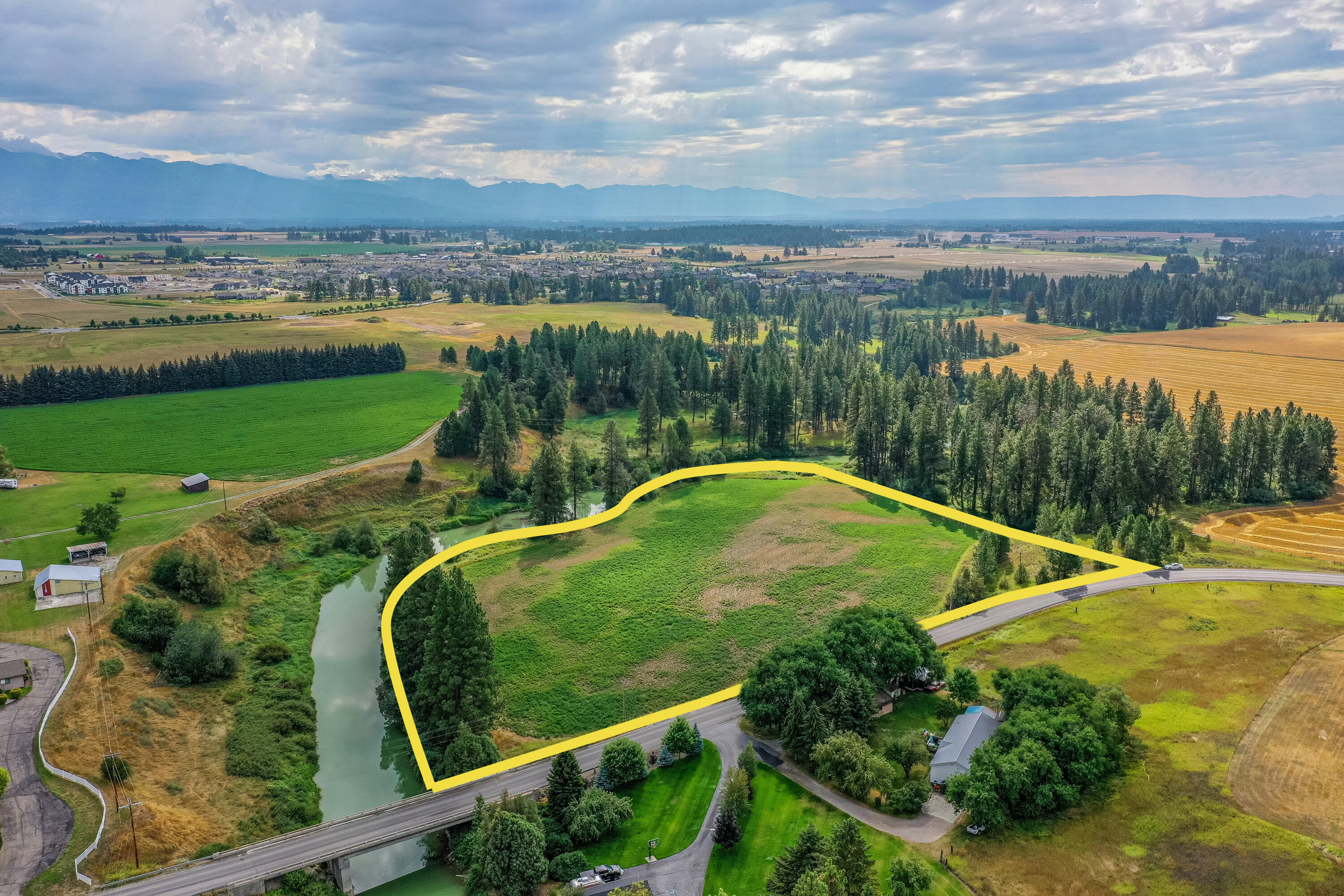 Aerial view of a green plot of land outlined in yellow, bordered by a river, trees, and a road with distant mountains.