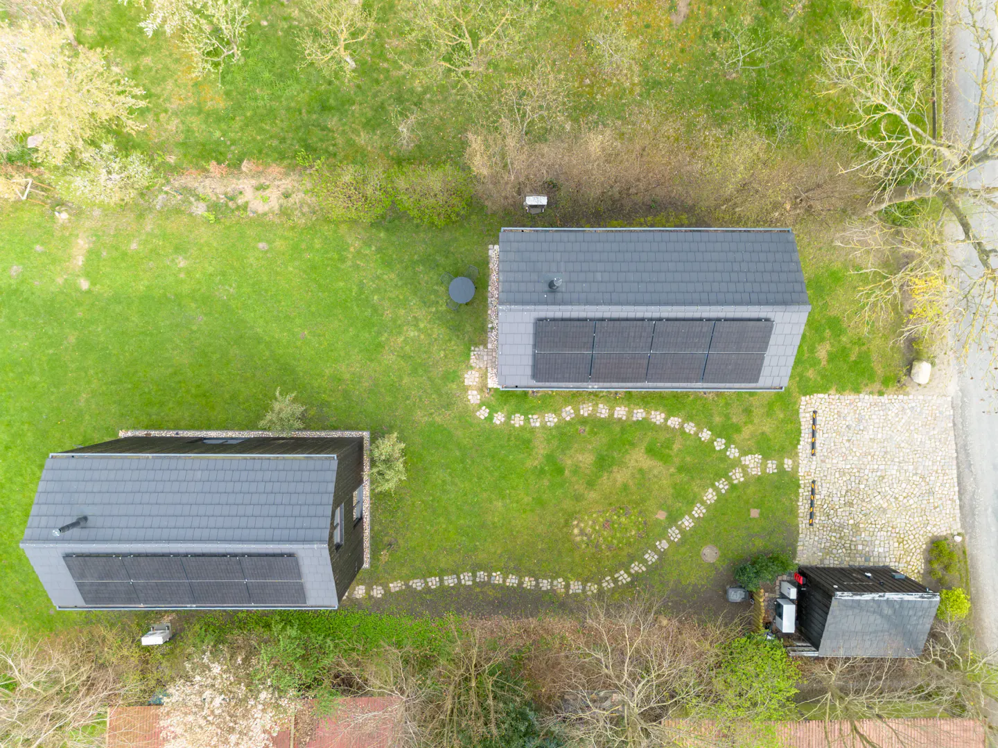 Aerial view of two modern houses with solar panels on gray roofs, connected by a stone path on green grass.