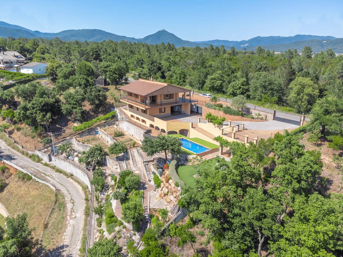 Aerial view of a two-story tan house with a brown roof, a pool, and lush green trees. Mountains are visible in the background.