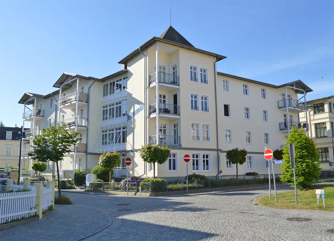 A three-story, light yellow apartment building with balconies and white trim on a cobblestone street.
