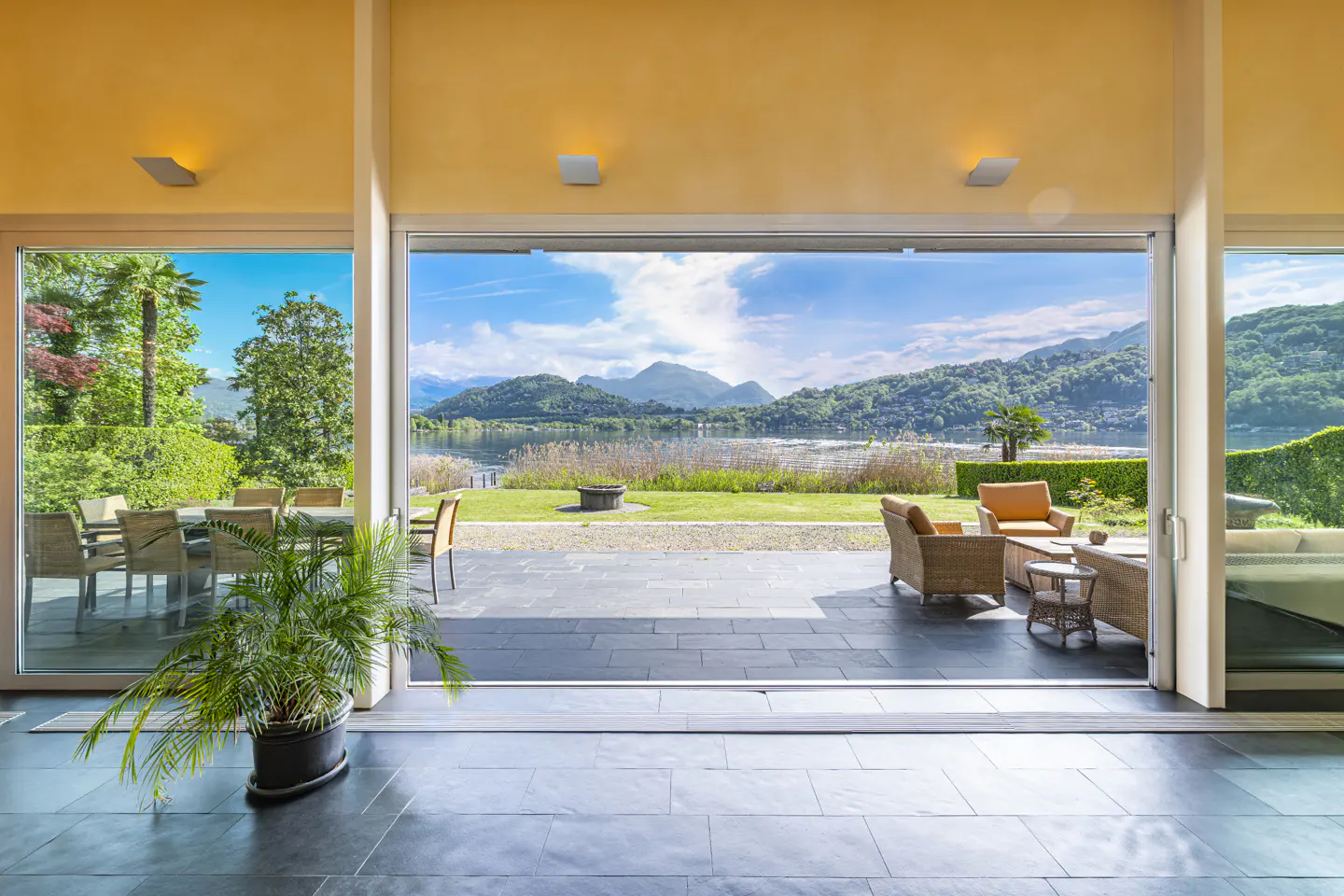 Open sliding glass doors reveal a patio with lake and mountain views. Outdoor seating and dining areas are visible.