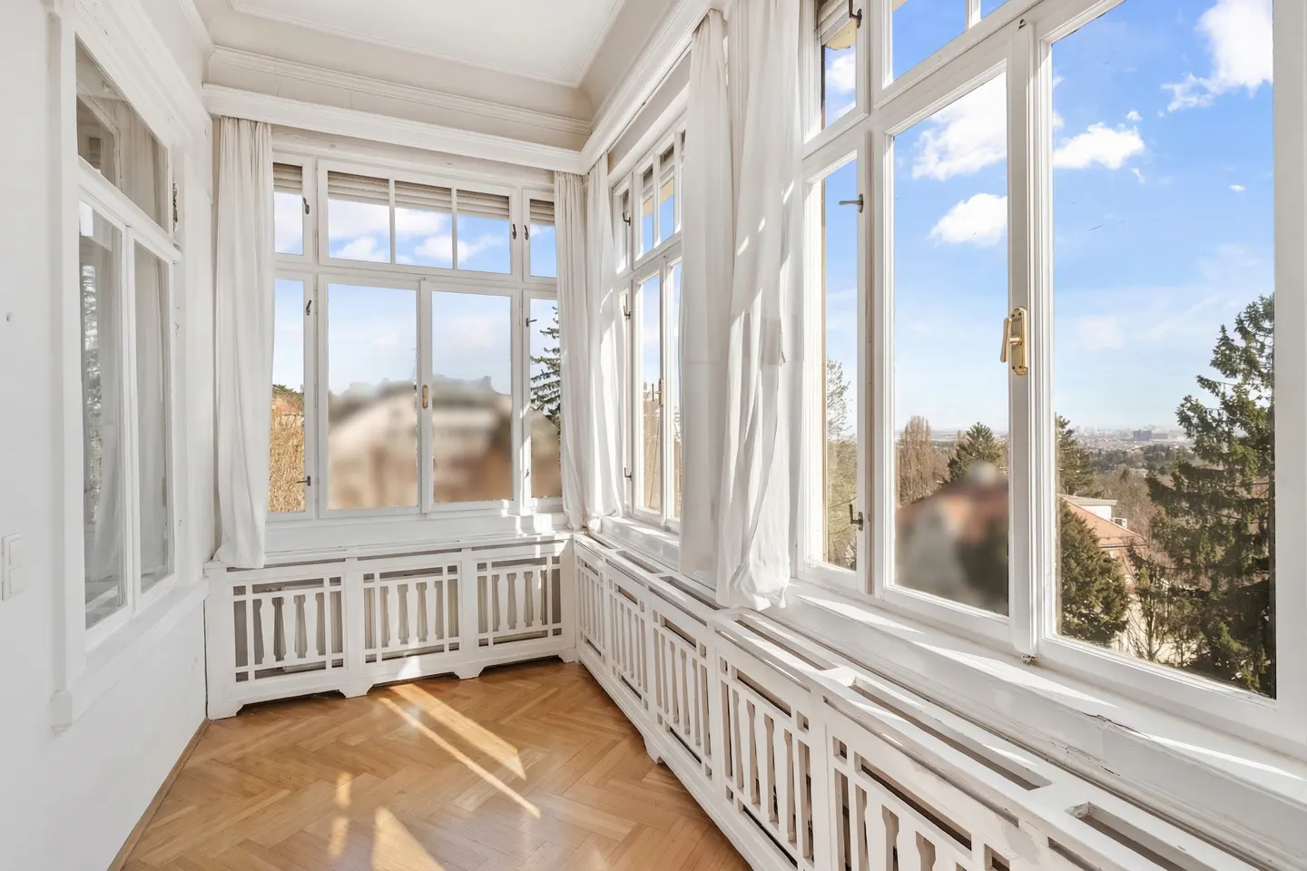 Bright sunroom with white trim, large windows, and herringbone wood floors. White curtains frame the windows with a view of trees and sky.