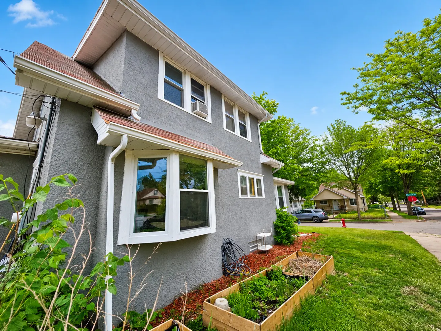 Gray two-story house with white trim, a bay window, and a red-tiled roof. A garden bed sits on the green lawn.