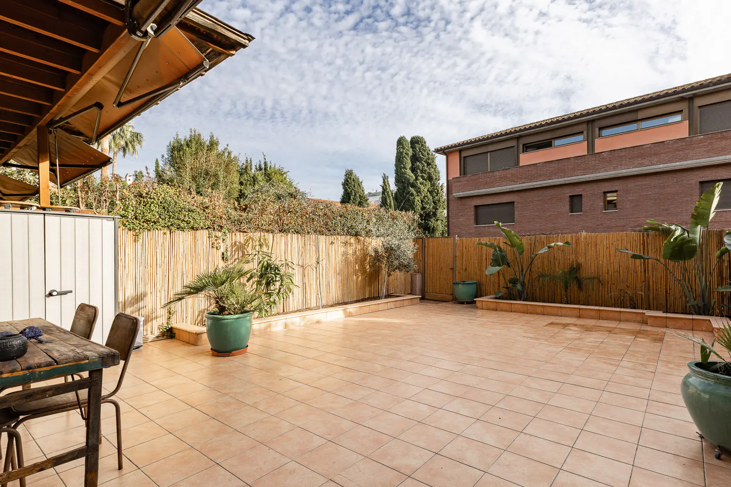 Outdoor patio with tile flooring, bamboo fence, and green potted plants. A wooden table and chairs sit under a partial awning.