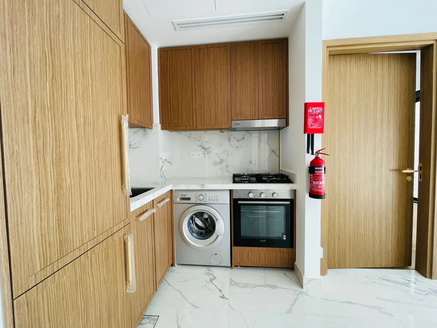 A modern kitchen with light wood cabinets, marble countertops, and a washing machine next to the oven. A red fire extinguisher is mounted near a wooden door.