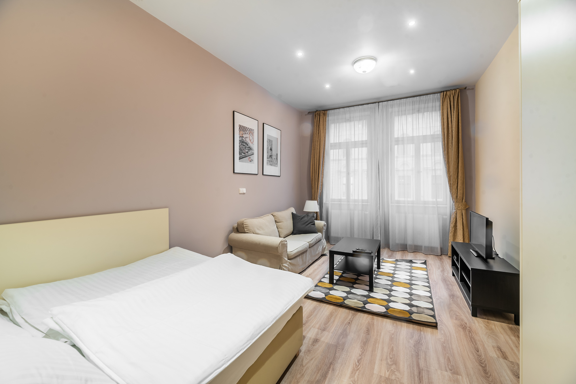 Studio apartment with a bed, beige sofa, black coffee table, patterned rug, and TV on a black stand near large windows. [AI-generated text]