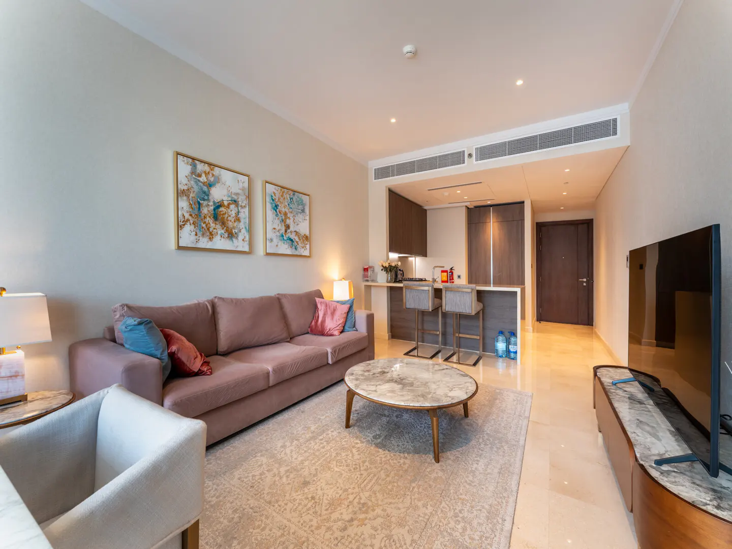 Living room with a pink couch, marble coffee table, and two abstract paintings on the wall. A kitchen bar with two stools is in the background.