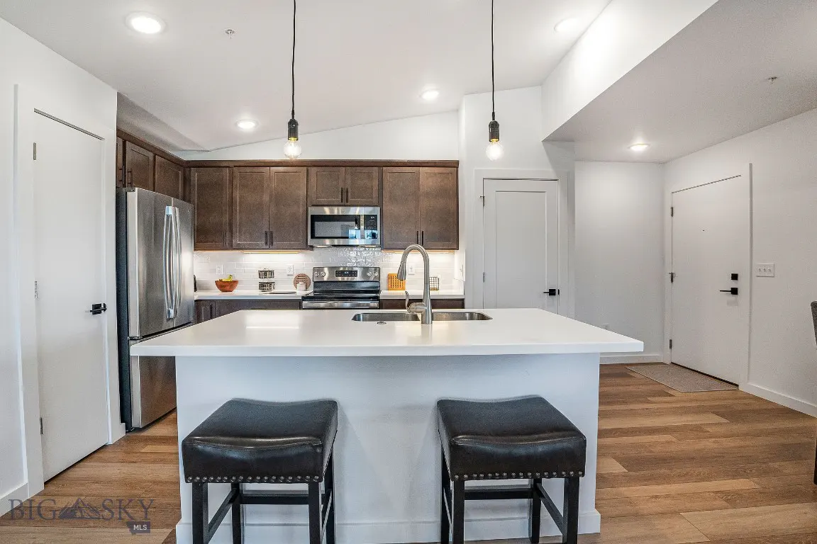 A modern kitchen with brown cabinets, stainless steel appliances, and a white countertop island with two black leather stools.