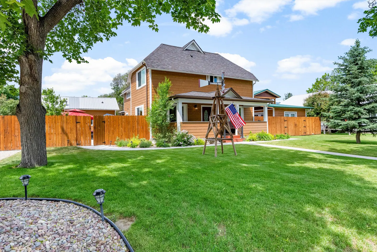Two-story brown house with a gray roof, white trim, and a wooden fence. A windmill with an American flag stands in the green lawn.