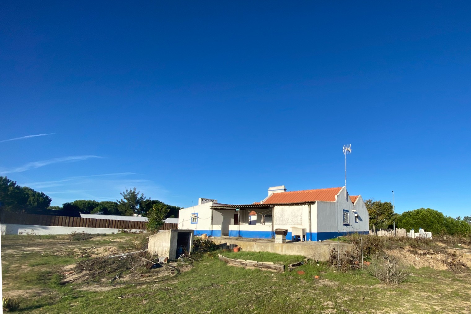 A white single-story house with a terracotta roof and blue trim sits on a grassy lot under a clear, bright blue sky. [AI-generated text]